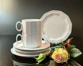 Pode incluir: Um conjunto de chávenas e pires de chá de porcelana branca com um padrão de listras azuis e cinzas. As chávenas estão empilhadas uma em cima da outra e os pires estão dispostos em círculo à sua volta. Uma única rosa amarela está sobre a mesa ao lado das chávenas.