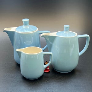 Può includere: Un set di tre teiere in ceramica blu chiaro con coperchi bianchi. La teiera a sinistra è più piccola di quella a destra. La teiera più piccola è un creamer.