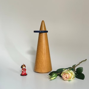 Może przedstawiać: Drewniany stojak na pierścionek w kształcie stożka z ciemnoniebieskim pierścionkiem na górze. Na pierwszym planie znajduje się mała czerwona figurka osoby. Na pierwszym planie znajduje się pojedyncza biała róża z zielonymi liśćmi.