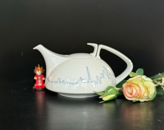 May include: White ceramic teapot with a blue cityscape design. The teapot has a round body and a spout on one side. The handle is on the opposite side of the spout. The teapot is sitting on a black surface.