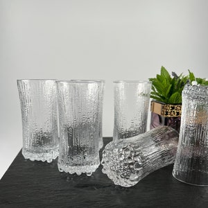 May include: A set of six clear, textured glass tumblers. The glasses have a vertical, bark-like pattern and a textured base. The glasses are arranged on a dark gray surface, with a small plant in the background.