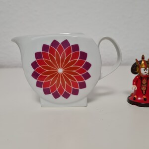 May include: White ceramic creamer with a red and purple floral design. A small red and gold figurine of a woman with a crown is in the background.