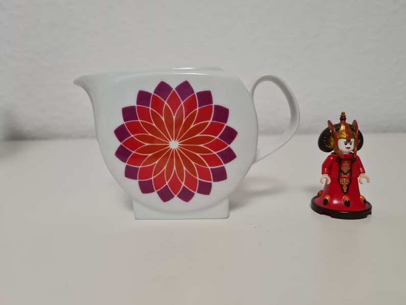 May include: White ceramic creamer with a red and purple floral design. A small red and gold figurine of a woman with a crown is in the background.