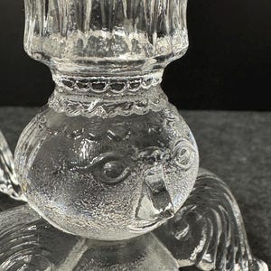 Puede incluir: Candelabro de &aacute;ngel de cristal transparente con rasgos faciales detallados, alas y una copa para velas. El candelabro est&aacute; dise&ntilde;ado con un cuerpo redondo y una superficie texturizada. Las alas del &aacute;ngel son visibles en el lateral.