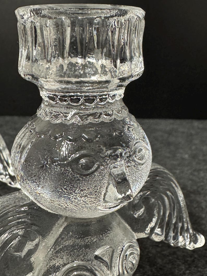 Puede incluir: Candelabro de &aacute;ngel de cristal transparente con rasgos faciales detallados, alas y una copa para velas. El candelabro est&aacute; dise&ntilde;ado con un cuerpo redondo y una superficie texturizada. Las alas del &aacute;ngel son visibles en el lateral.