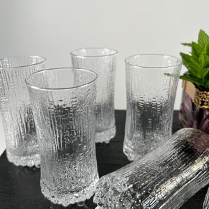 May include: A set of five clear, textured drinking glasses. The glasses have a cylindrical shape with a textured, bark-like pattern. One glass is lying on its side. The glasses are on a dark surface.