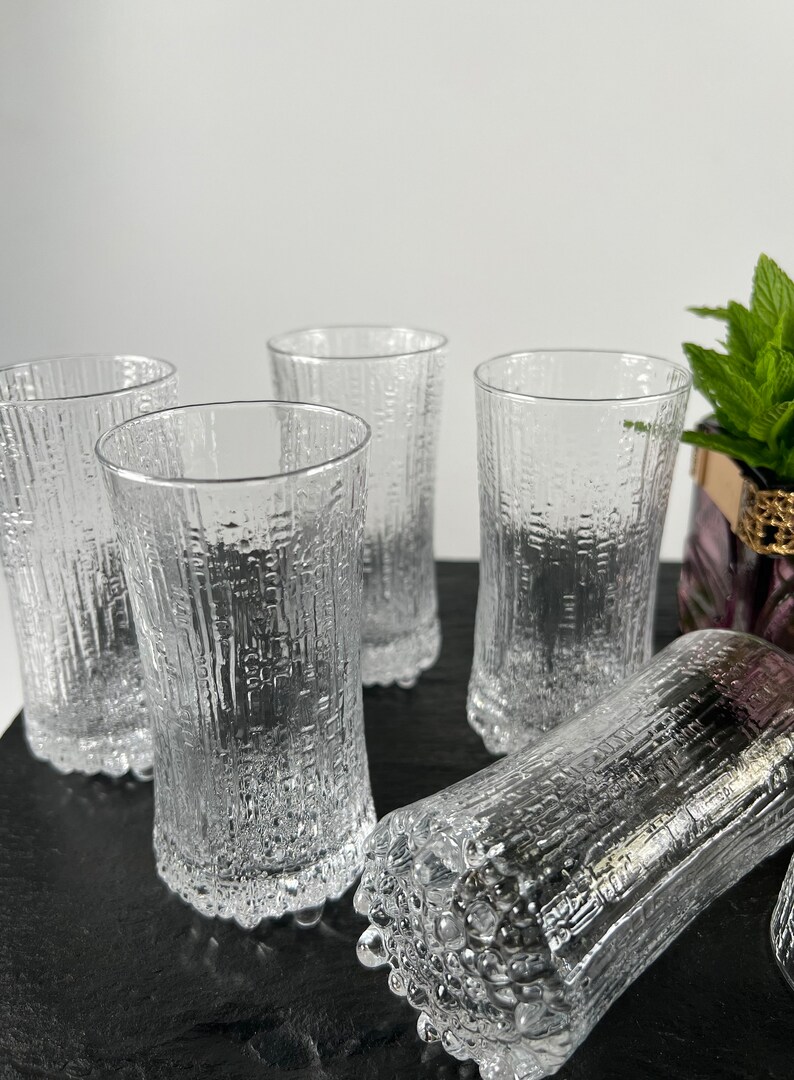 May include: A set of five clear, textured drinking glasses. The glasses have a cylindrical shape with a textured, bark-like pattern. One glass is lying on its side. The glasses are on a dark surface.