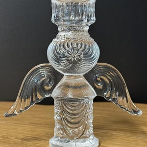Puede incluir: Un portavelas transparente con forma de &aacute;ngel de cristal. El portavelas tiene un dise&ntilde;o detallado con alas, una secci&oacute;n media redonda y una base. Las alas est&aacute;n extendidas hacia los lados. El portavelas est&aacute; sobre una superficie de madera.