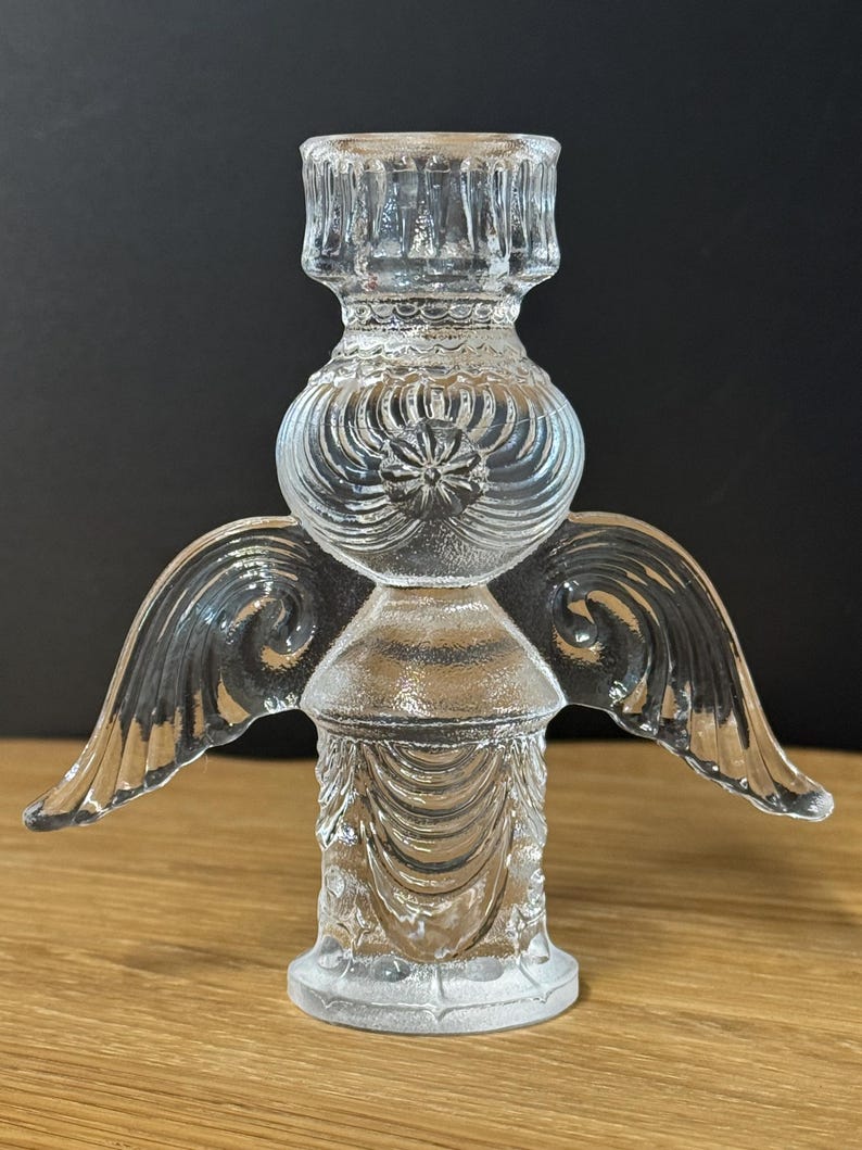 Puede incluir: Un portavelas transparente con forma de &aacute;ngel de cristal. El portavelas tiene un dise&ntilde;o detallado con alas, una secci&oacute;n media redonda y una base. Las alas est&aacute;n extendidas hacia los lados. El portavelas est&aacute; sobre una superficie de madera.