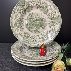 Könnte beinhalten: Set aus vier grünen und weißen, floral gemusterten Keramik-Dinnertellern. Die Teller haben einen gewellten Rand und zeigen eine Cottage-Szene mit einer Brücke und Bäumen.