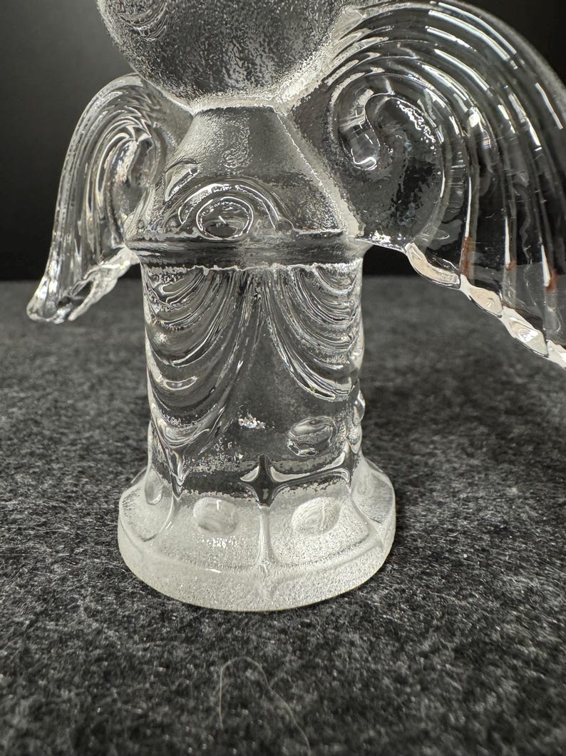 Puede incluir: Una figura de &aacute;ngel de cristal transparente con dise&ntilde;os detallados de alas y cuerpo. El &aacute;ngel tiene una superficie texturizada y est&aacute; sobre una base redonda. Las alas est&aacute;n extendidas y el cuerpo tiene un patr&oacute;n decorativo.