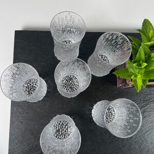 May include: Six clear textured drinking glasses arranged on a black surface. The glasses have a unique pattern and a textured design. The glasses are empty and ready to be filled with a beverage.
