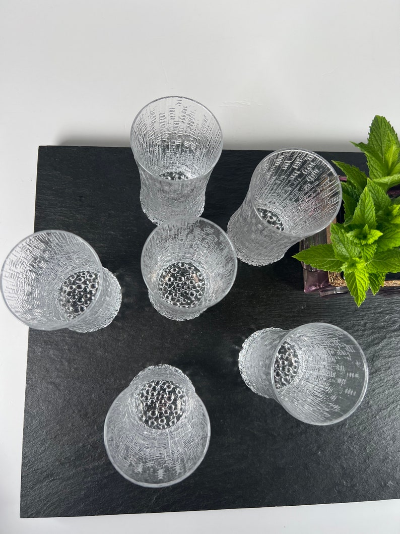 May include: Six clear textured drinking glasses arranged on a black surface. The glasses have a unique pattern and a textured design. The glasses are empty and ready to be filled with a beverage.