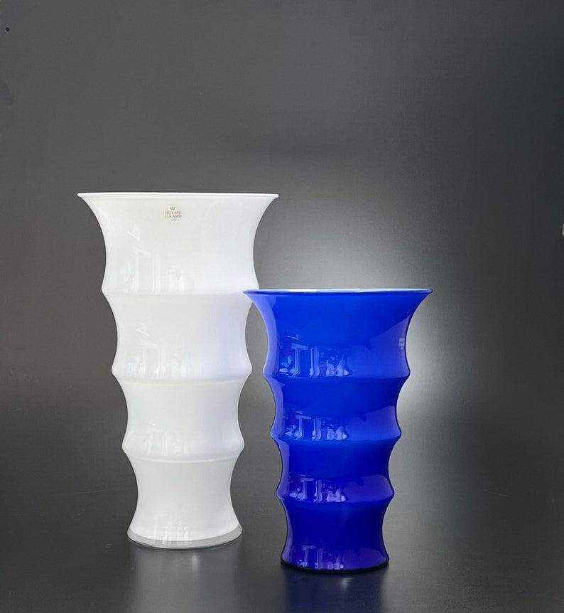 May include: Two glass vases, one white and one blue, with a textured, ribbed design. The white vase has the text "Kosta Boda" on the side.