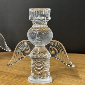 Puede incluir: Candelabro de &aacute;ngel de cristal transparente con alas detalladas y una cara sonriente. El candelabro tiene una base redonda y una parte superior cil&iacute;ndrica para sostener una vela. La figura del &aacute;ngel es de cristal transparente.