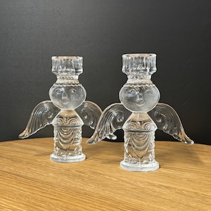 Puede incluir: Dos candelabros de &aacute;ngel de cristal transparente. Cada uno tiene una cabeza redonda con una cara, alas y una base detallada. Los candelabros est&aacute;n sobre una superficie de madera con un fondo oscuro.