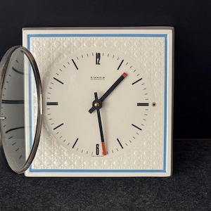 Könnte beinhalten: Quadratischer weißer Kienzle Wecker mit hellblauem Rand und strukturierter Oberfläche. Das Zifferblatt ist weiß mit schwarzen Zeigern und Ziffern sowie einem roten Akzent am Minutenzeiger. Die Uhr hat einen silberfarbenen Klappdeckel.