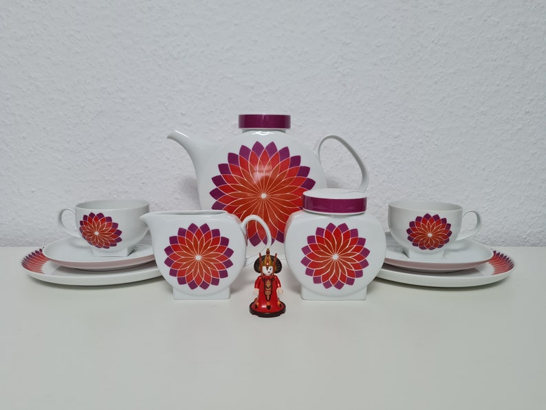 May include: A white tea set with a red and purple floral pattern. The set includes a teapot, creamer, sugar bowl, two cups, and two saucers. A small red figurine is in the center of the image.