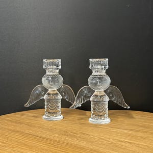 Puede incluir: Dos candelabros de &aacute;ngel de cristal transparente. Cada &aacute;ngel tiene alas, una cabeza redonda con una cara sonriente y un cuerpo detallado. Los candelabros est&aacute;n sobre una superficie de madera contra un fondo oscuro.