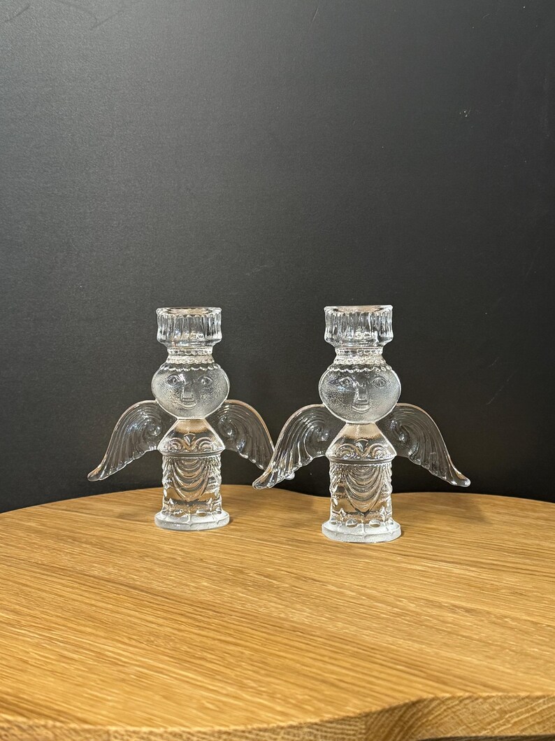 Puede incluir: Dos candelabros de &aacute;ngel de cristal transparente. Cada &aacute;ngel tiene alas, una cabeza redonda con una cara sonriente y un cuerpo detallado. Los candelabros est&aacute;n sobre una superficie de madera contra un fondo oscuro.