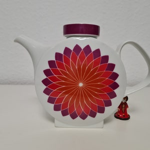 May include: White teapot with a round body and a purple lid. The teapot has a geometric design of red and purple petals on the front.
