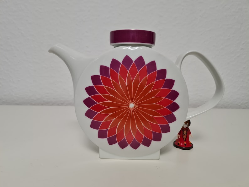 May include: White teapot with a round body and a purple lid. The teapot has a geometric design of red and purple petals on the front.