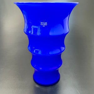 May include: A cobalt blue glass vase with a flared top and a rippled design. The vase is marked with the text "Holme Gaard 1823".