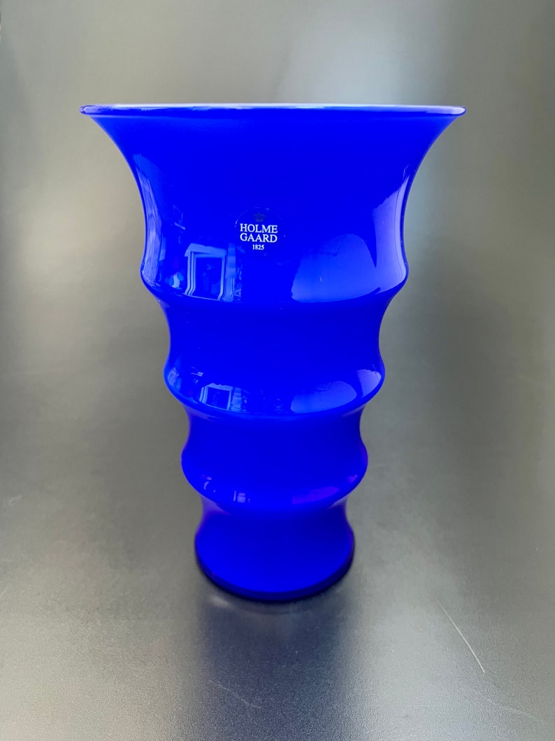 May include: A cobalt blue glass vase with a flared top and a rippled design. The vase is marked with the text "Holme Gaard 1823".