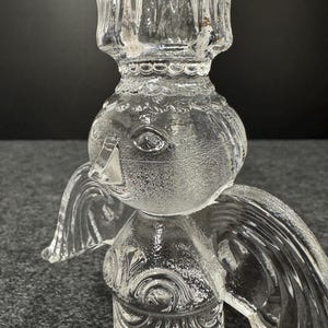 Puede incluir: Candelabro de vidrio transparente con forma de &aacute;ngel. El candelabro tiene un dise&ntilde;o detallado con alas y una cabeza redonda. El candelabro est&aacute; hecho de vidrio transparente y tiene una superficie texturizada.