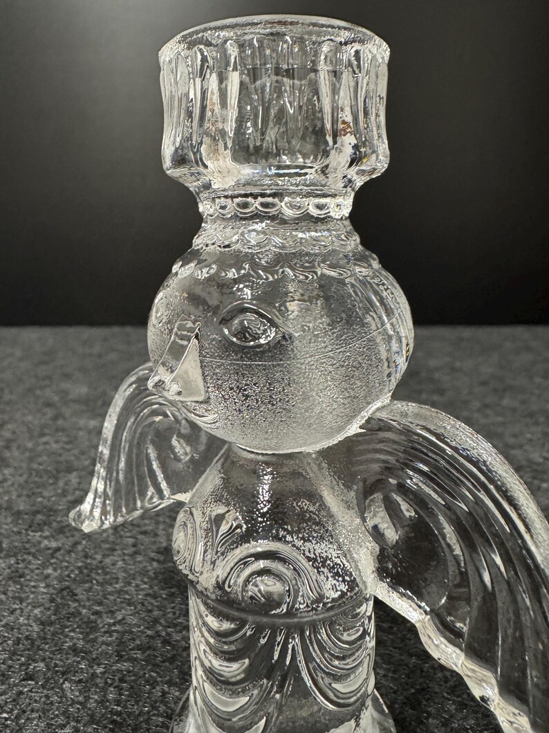 Puede incluir: Candelabro de vidrio transparente con forma de &aacute;ngel. El candelabro tiene un dise&ntilde;o detallado con alas y una cabeza redonda. El candelabro est&aacute; hecho de vidrio transparente y tiene una superficie texturizada.