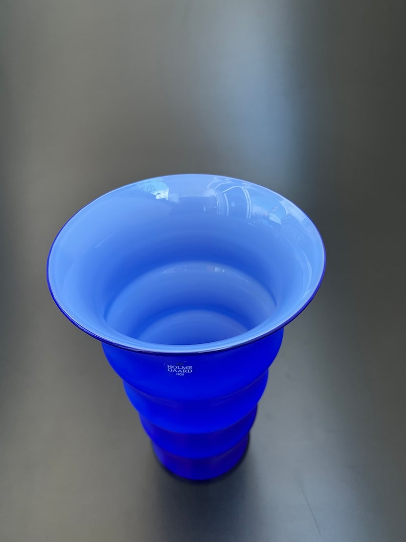 May include: A blue glass vase with a white interior and a rippled design. The vase is marked with the text "Holme Gaard 1825".
