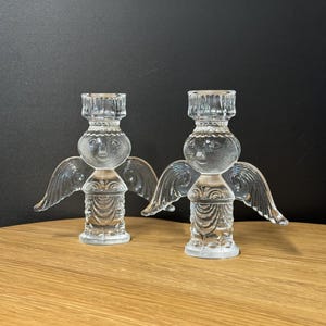 Puede incluir: Dos candelabros de &aacute;ngel de cristal transparente. Cada &aacute;ngel tiene una cabeza redonda con una cara, alas y un cuerpo detallado. Los candelabros est&aacute;n sobre una superficie de madera contra un fondo oscuro.