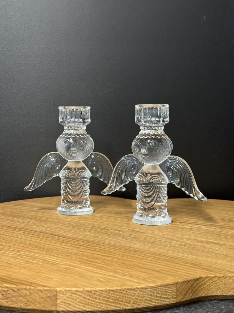 Puede incluir: Dos candelabros de &aacute;ngel de cristal transparente. Cada &aacute;ngel tiene una cabeza redonda con una cara, alas y un cuerpo detallado. Los candelabros est&aacute;n sobre una superficie de madera contra un fondo oscuro.