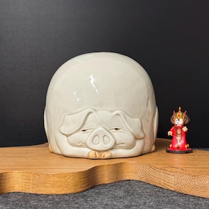 Könnte beinhalten: Ein großer, cremefarbener Keramikbehälter in Schweineform mit detaillierter Schnauze und geschlossenen Augen. Rechts steht eine kleine Figur einer Person in roter und goldener Kleidung. Die Objekte stehen auf einer Holzoberfläche.