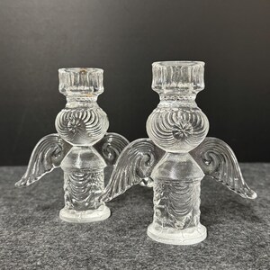Puede incluir: Dos candelabros de &aacute;ngel de cristal transparente. Cada candelabro tiene una base redonda, un cuerpo cil&iacute;ndrico y una parte superior dise&ntilde;ada para sostener una vela. Los &aacute;ngeles tienen alas detalladas y se exhiben sobre una superficie gris.
