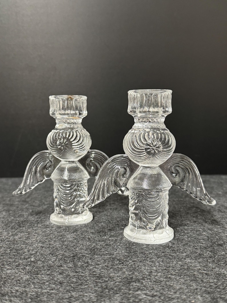 Puede incluir: Dos candelabros de &aacute;ngel de cristal transparente. Cada candelabro tiene una base redonda, un cuerpo cil&iacute;ndrico y una parte superior dise&ntilde;ada para sostener una vela. Los &aacute;ngeles tienen alas detalladas y se exhiben sobre una superficie gris.