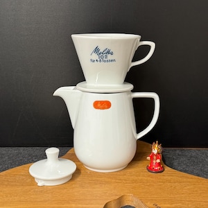 May include: White ceramic Melitta coffee maker set with a pour-over filter, a carafe, and a lid. The filter is labeled "Melitta 102 für 4-8 Tassen." A small figurine sits on a wooden serving board.