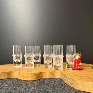 May include: Six clear glass shot glasses with a textured surface are arranged in a row on a wooden chopping board. A small red figurine is to the right of the glasses.