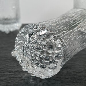 May include: Close-up of a clear glass tumbler with a textured, bubble-like surface. The cylindrical glass has a unique design, with a rough, uneven texture. The glass is partially in focus, with a blurred background.
