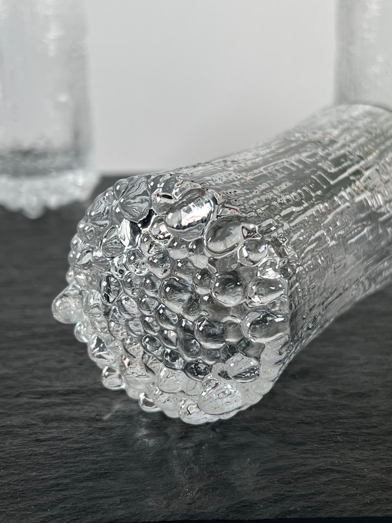 May include: Close-up of a clear glass tumbler with a textured, bubble-like surface. The cylindrical glass has a unique design, with a rough, uneven texture. The glass is partially in focus, with a blurred background.