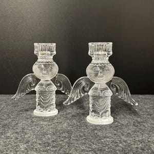 Puede incluir: Dos candelabros de &aacute;ngel de cristal transparente. Cada &aacute;ngel tiene una cara, alas y un cuerpo detallado. Los candelabros est&aacute;n dise&ntilde;ados para sostener una sola vela. El fondo es una superficie gris oscuro.