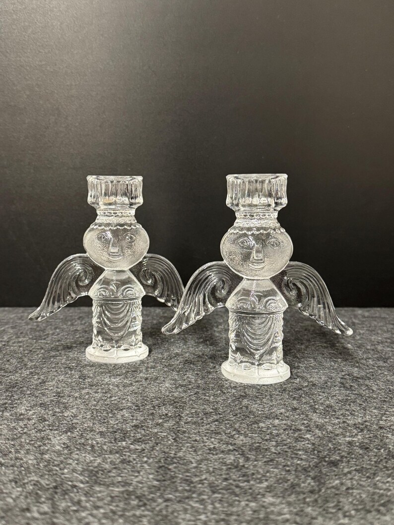 Puede incluir: Dos candelabros de &aacute;ngel de cristal transparente. Cada &aacute;ngel tiene una cara, alas y un cuerpo detallado. Los candelabros est&aacute;n dise&ntilde;ados para sostener una sola vela. El fondo es una superficie gris oscuro.
