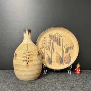 May include: A beige ceramic vase and plate set with brown and black floral designs. The vase has a narrow neck and bulbous body. The plate is round and rests on a clear stand. A small red figurine is also present.