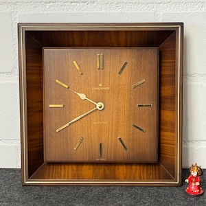 May include: A square, wooden wall clock with a brown finish and gold-colored accents. The clock face features the brand name "Junghans" and the word "quartz". The clock has gold-colored hands and markers. A small decorative figure is in the lower right corner.