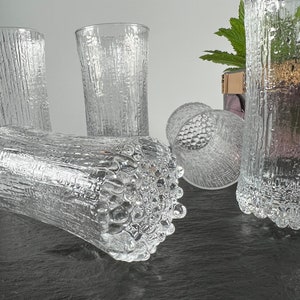 May include: Clear textured glass tumblers. The glasses have a bark-like texture on the exterior. One tumbler is lying on its side, while others stand upright. The glassware is suitable for water, juice, or other beverages.