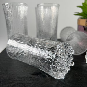 May include: Clear glass tumblers with a textured, bark-like design. The glasses are cylindrical and appear to be made of thick glass. Several glasses are visible, some upright and others lying on their side. The background is a neutral color.