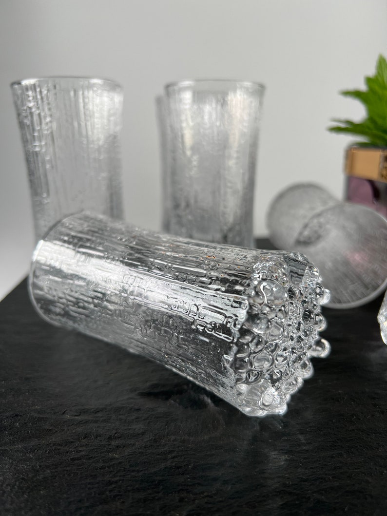May include: Clear glass tumblers with a textured, bark-like design. The glasses are cylindrical and appear to be made of thick glass. Several glasses are visible, some upright and others lying on their side. The background is a neutral color.