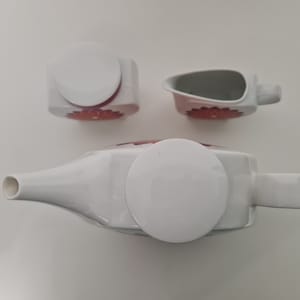 May include: A white ceramic teapot, creamer, and sugar bowl set with red floral designs. The teapot and creamer have lids.