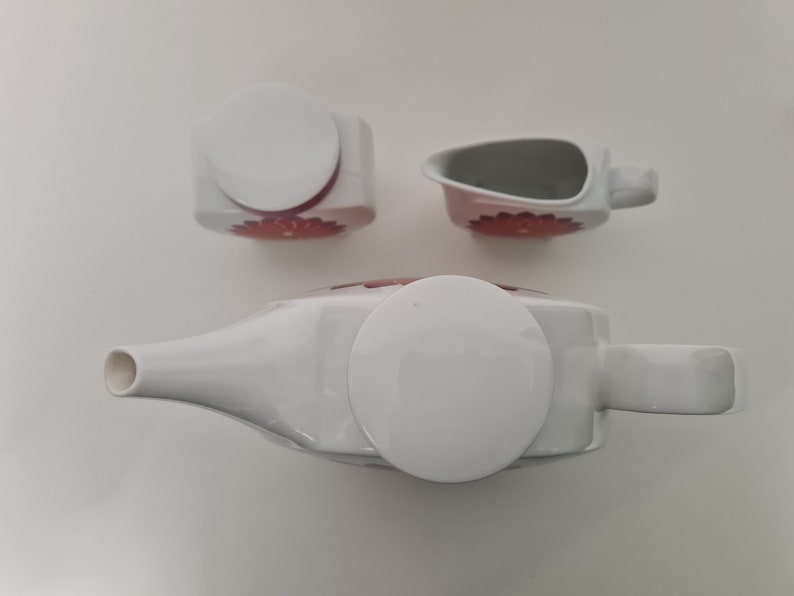 May include: A white ceramic teapot, creamer, and sugar bowl set with red floral designs. The teapot and creamer have lids.