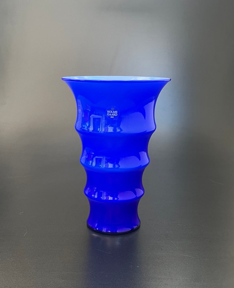 May include: A cobalt blue glass vase with a flared top and a rippled design. The vase is marked with the text "Holme Gard" in white.